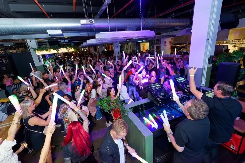 A packed Auckland dancefloor with illuminated foam batons beside the DJ booth